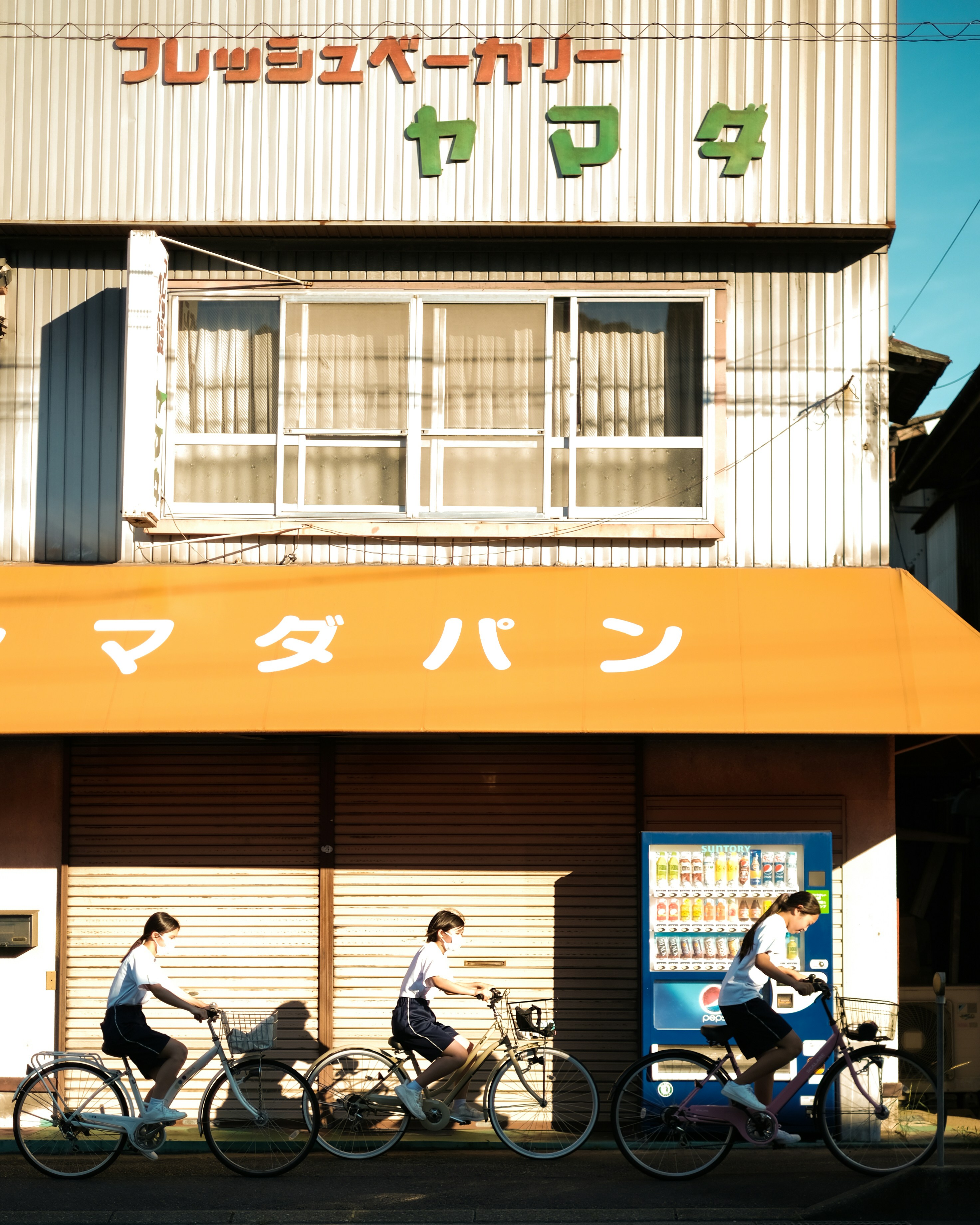 Three students riding bicycles past a Japanese bakery storefront